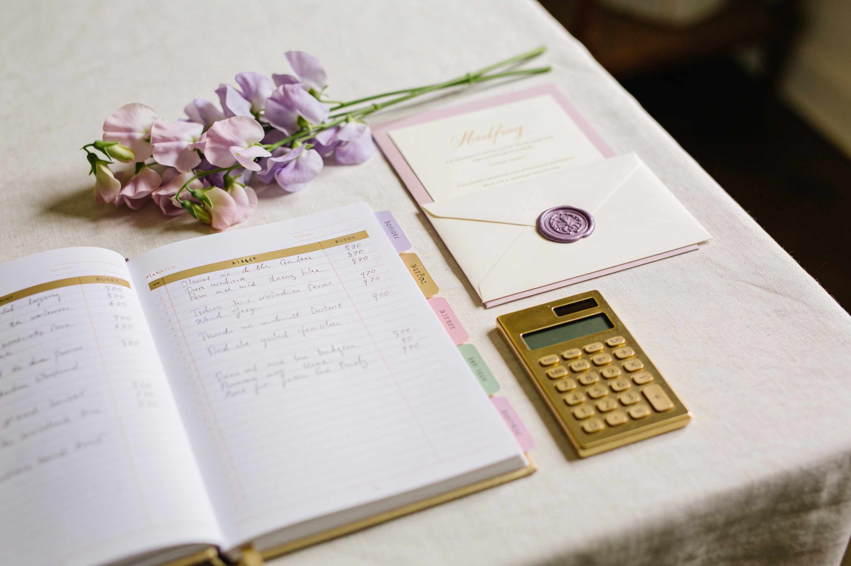 A planning notebook open to a handwritten budget page with pastel tabs, a gold calculator, an invitation with a lilac wax seal and sweet peas.