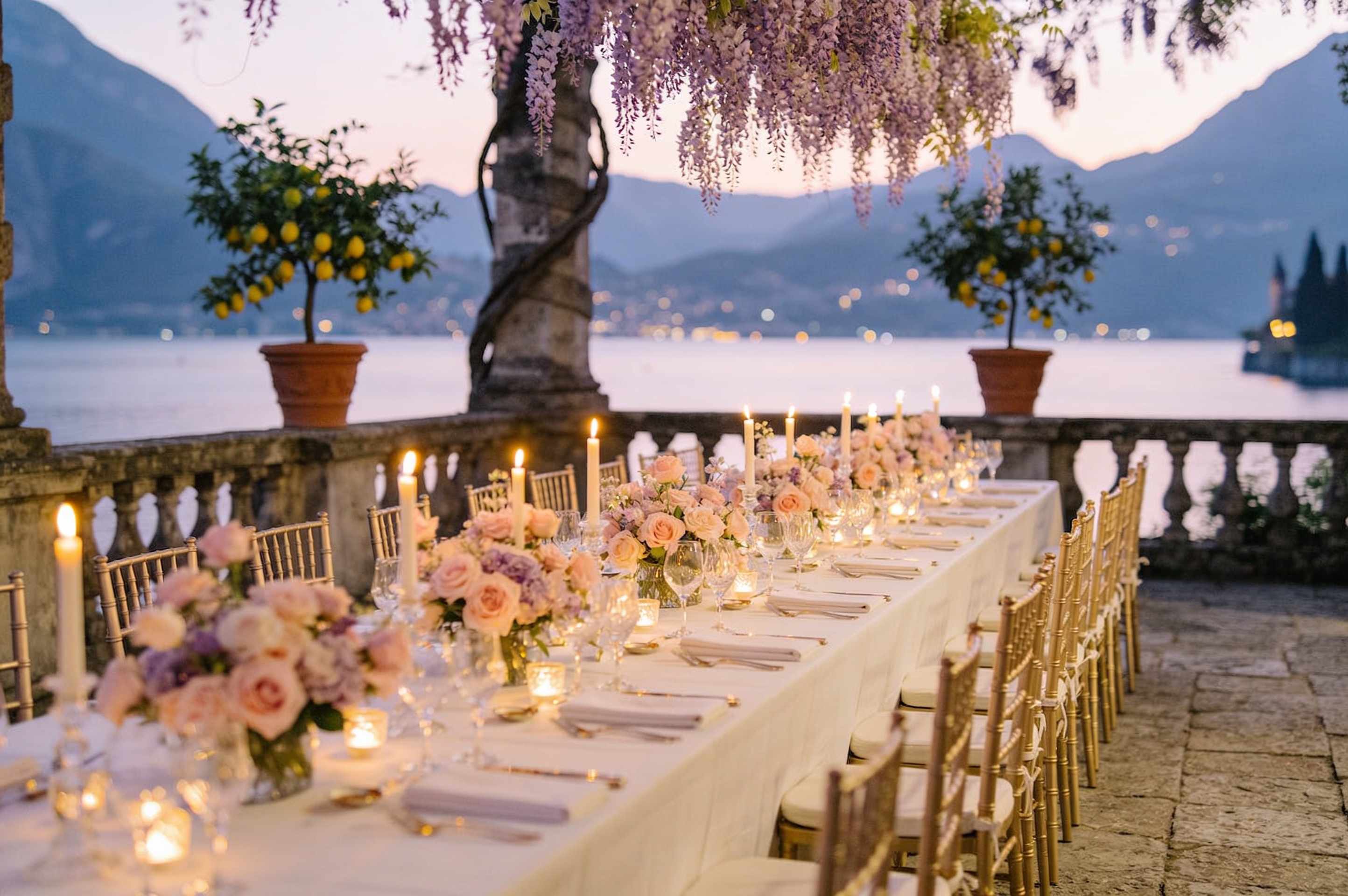 A candlelit reception table on the terrace of a lakeside villa at dusk, with lemon trees, blush roses and wisteria, mountains glowing lavender beyond.