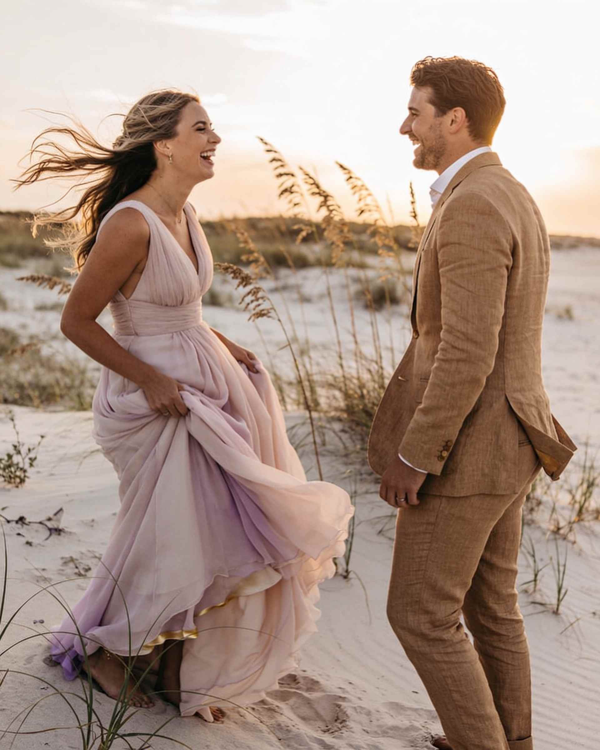 A bride in a blush chiffon gown and a groom in a tan linen suit laughing together on a windswept beach among sea oats at sunset.
