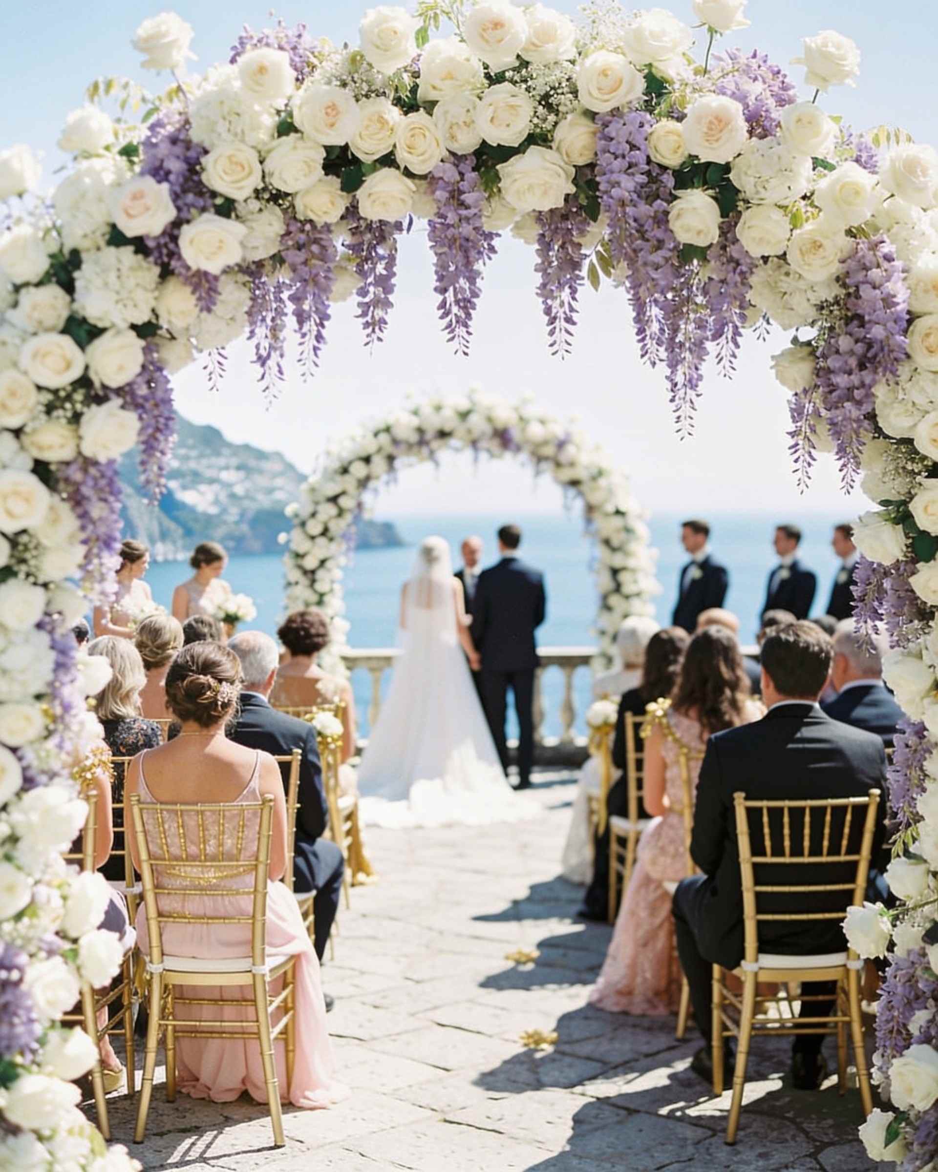 A ceremony on a stone terrace above the sea with an arch of white roses and wisteria, guests seated in rows and the couple at the arch.