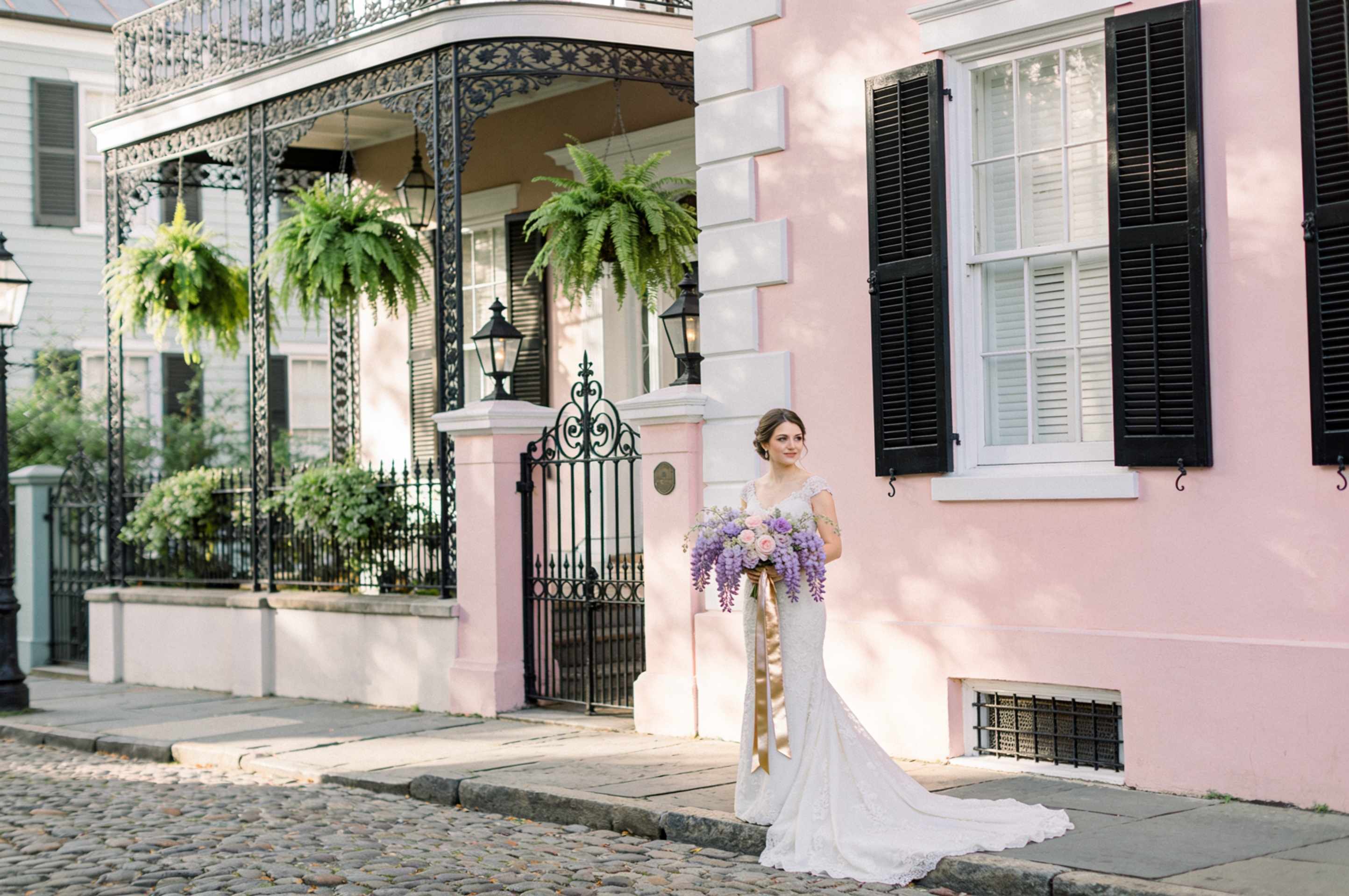 A blush pink Charleston single house with black shutters, a piazza porch with hanging ferns and a wrought iron gate, a bride in white on the cobblestones outside.